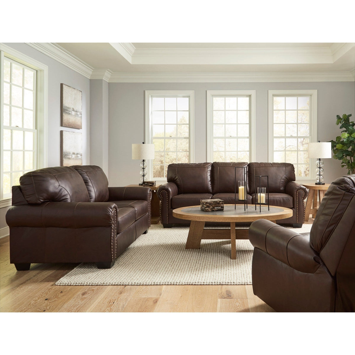 A full living room scene showcasing the dark brown loveseat, sofa, and recliner in a bright, traditionally styled space with light gray walls and large windows.