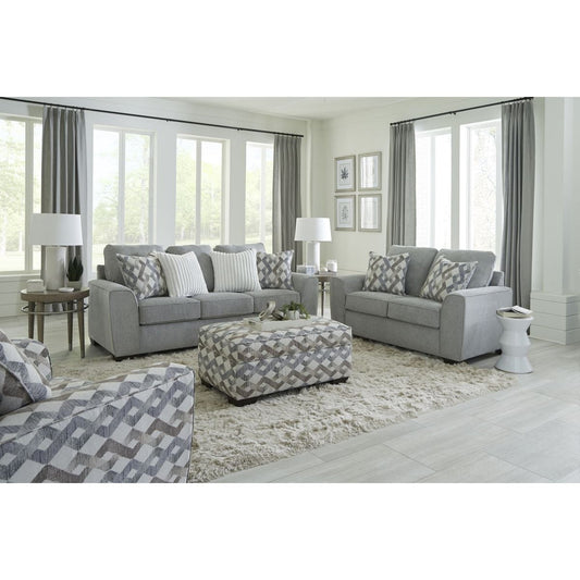The steel gray loveseat is showcased as part of a complete living room set, featuring its clean lines and two decorative geometric pillows, set in a bright, modern living space.